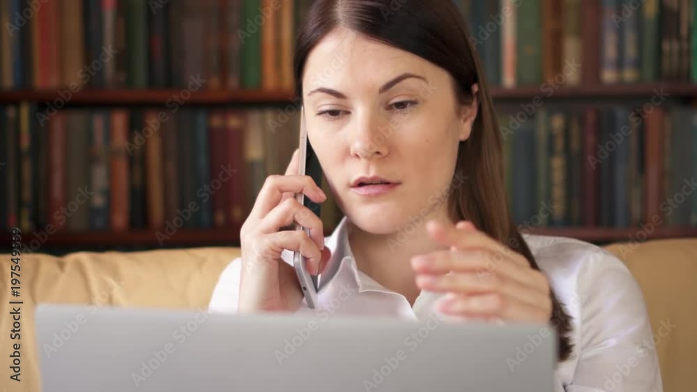 Busy businesswoman in white shirt at home office with laptop and smartphone. Professional female using cellphone discussing project on screen. Remote freelance work. Home-office concept