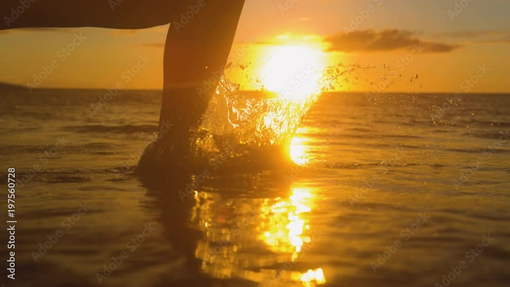 SLOW MOTION, CLOSE UP, LENS FLARE: Carefree woman runs barefoot in shallow ocean water at golden sunset. Young female traveler unwinds by running and splashing in the sea on beautiful summer morning.