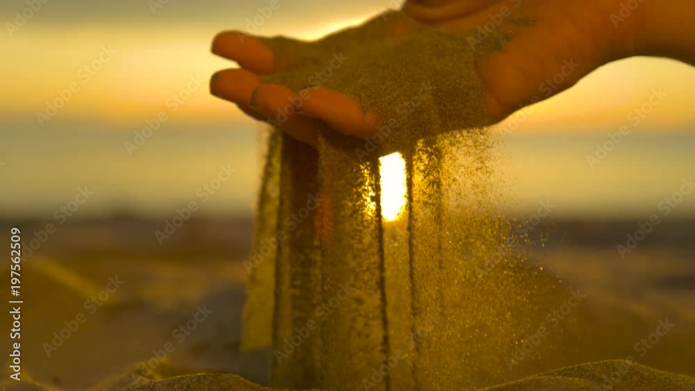 SLOW MOTION CLOSE UP, REVERSED: Small particles of dry sand fall between female fingers and back on shore. Beautiful summer sunset illuminates sandy beach and woman sifting dry sand with her hands.