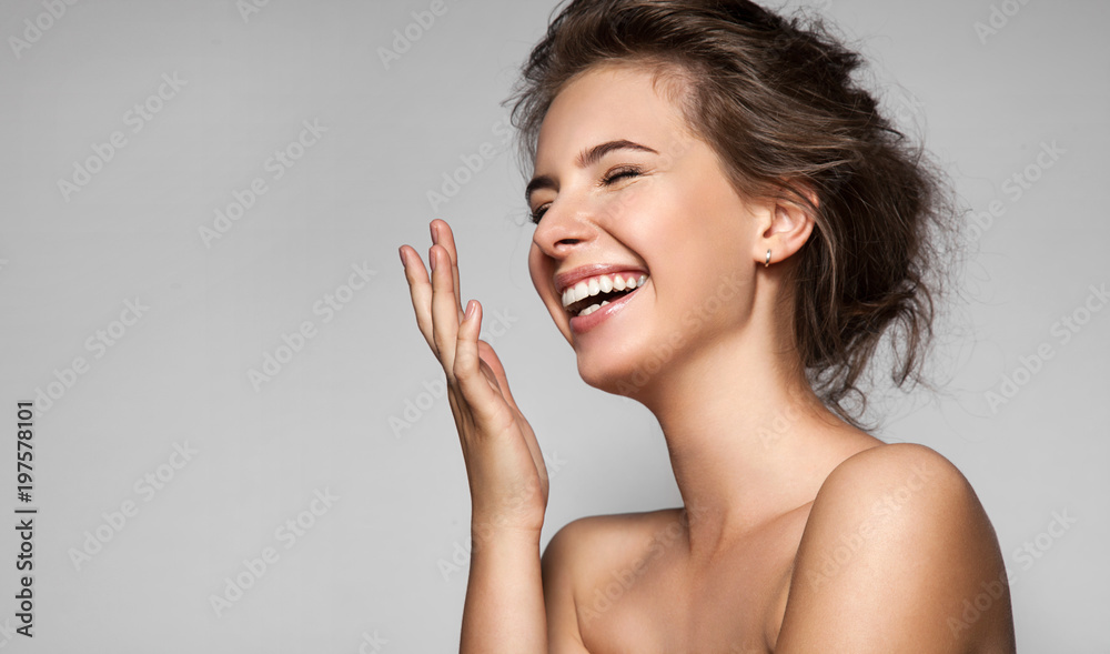 Naklejka premium Szczęśliwa roześmiana młoda kobieta o doskonałej skórze, naturalnym makijażu i pięknym uśmiechu. Żeński portret z nagimi ramionami na szarym tle