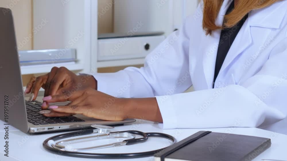 unrecognizable doctor sitting at the workspace working on laptop. african woman wearing white lab coat on the desk stethoscope. hospital details unrecognizable doctor sitting at the workspace working on laptop. african woman wearing white lab coat on the desk stethoscope. hospital details