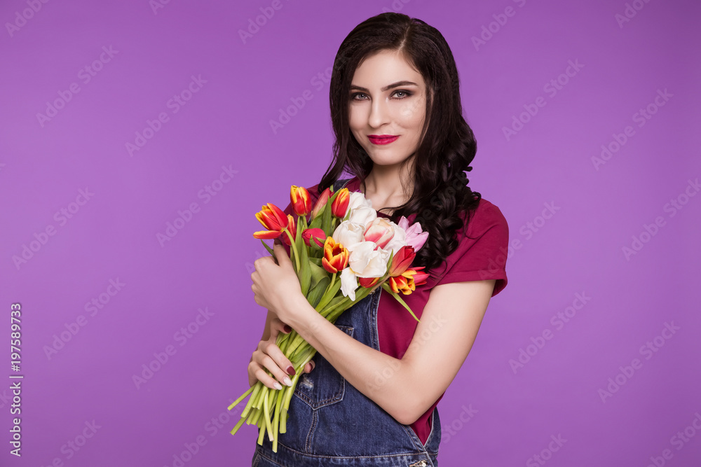 Beautiful woman, brunette, stands on purple background, in denim overalls and T-shirt with bouquet of tulips. The feminist. International Women's Day, March 8.