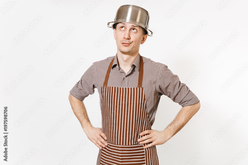 Fun man in striped apron with silver stainless glossy aluminium empty stewpan, pan or pot on head isolated on white background. Male housekeeper or houseworker. Kitchenware, dishes, cuisine concept.