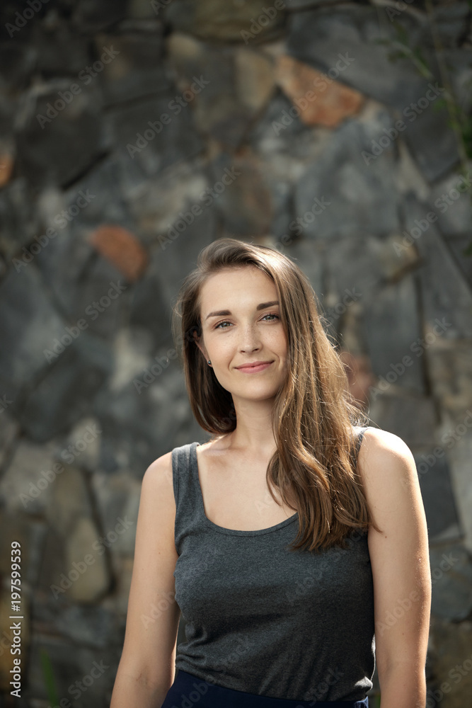 Waist-up portrait of naturally beautiful young woman smiling at camera on stone background