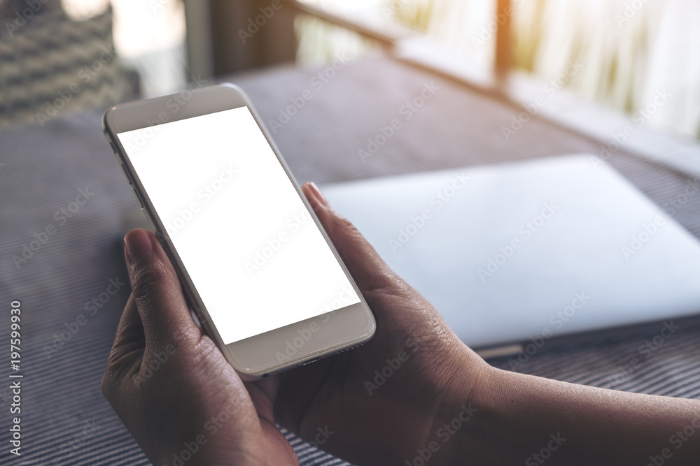 Mockup image of hands holding white mobile phone with blank desktop screen and laptop on table