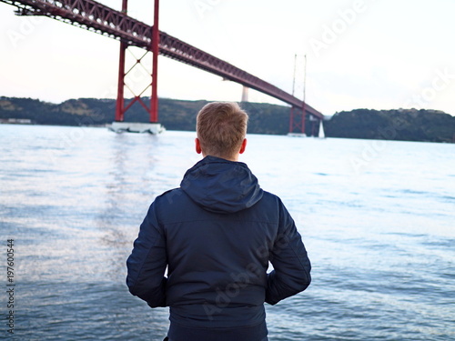 Photography A man in a jacket stands against the background of the sea city and the bridge in  Lisbon