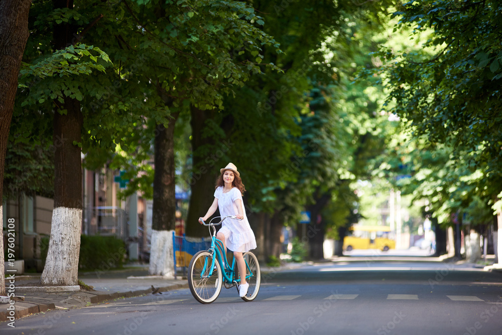 Summer sunny day. A nice girl on a bike alone on the road. Juicy city greens, vintage blue bicycle, a girl dressed in a light dress and hat