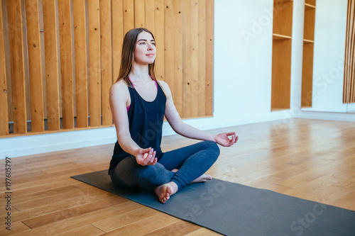 European woman practicing yoga, sitting in lotus pose, working out, wearing black t-shirt, pants, meditation session at home or modern studio interior. Full length photo