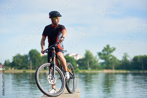 Sporty guy cyclist in cycling clothes and protective helmet posing near bicycle looking at side thinking about future success and achievement. Sportsman taking break at pier near lake.