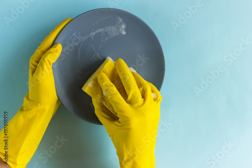 The housewife in yellow rubber gloves washes the plate with a yellow sponge. Blue background. Place for text. Flat lay. Concept