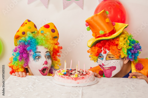 Clowns are a boy and a girl in bright costumes at the child s birthday. A table with refreshments and a cake. The explosion of emotions and the fun of the circus. greed and desire to eat cake