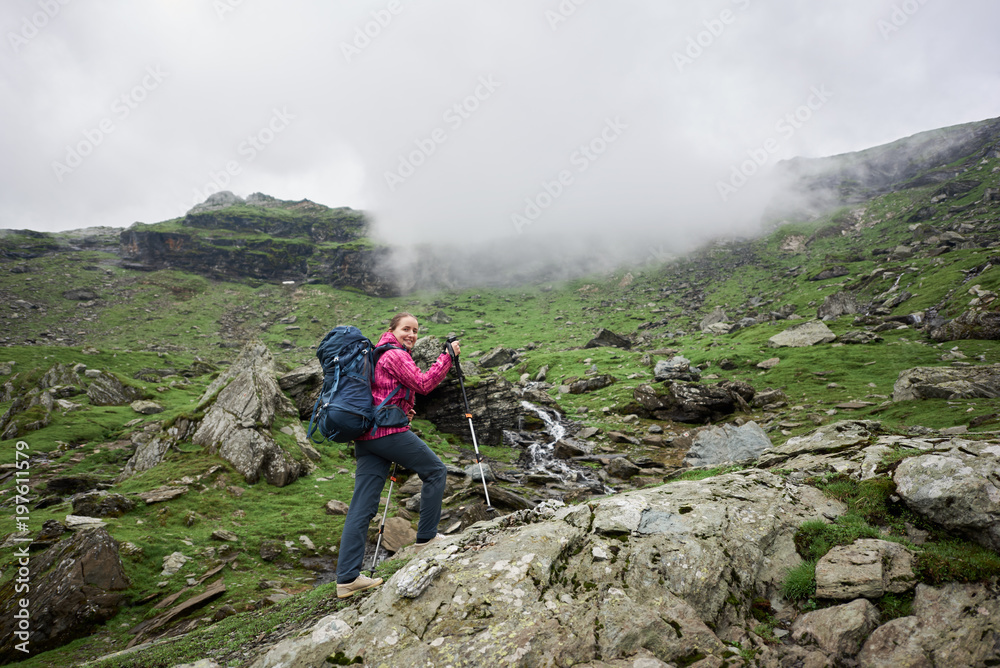 Positive female tourist climbing rock with walking sticks in front of green rocky meadows and mountains in Romania. Woman climber beautiful breathtaking view fog top