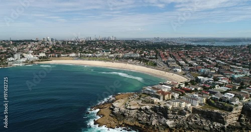 Rocky north bondi head approaching from open sea towards sandy stripe of beach with distant Sydney city CBD landmarks.
 photo