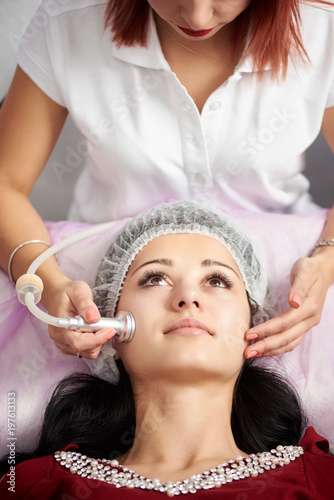 Brunette woman receiving electric ultrusound facial massage at beauty salon. Procedure in beauty clinic. Positive effects of physiotherapy. Close-up
