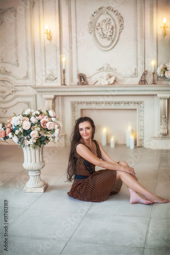 Lovely girl in brown dress sitting on floor near blooming flowers in luxurious room