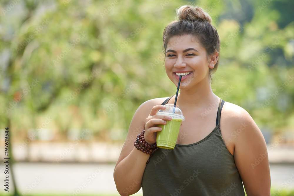Smiling young woman wearing top looking at camera while drinking smoothie from straw and enjoying picturesque view at green public park Smiling young woman wearing top looking at camera while drinking smoothie from straw and enjoying picturesque view at green public park