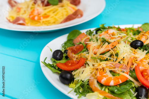 Close up of delicious salad in white plate on blue wooden table, spaghetti in the background