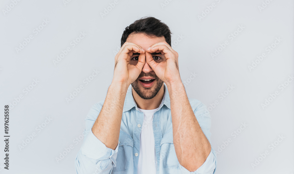 Handsome amazed male wearing blue shirt and white T-shirt holding his hands at his eyes as if looking through binoculars or glasses, surprised happily against studio light wall background. People Handsome amazed male wearing blue shirt and white T-shirt holding his hands at his eyes as if looking through binoculars or glasses, surprised happily against studio light wall background. People