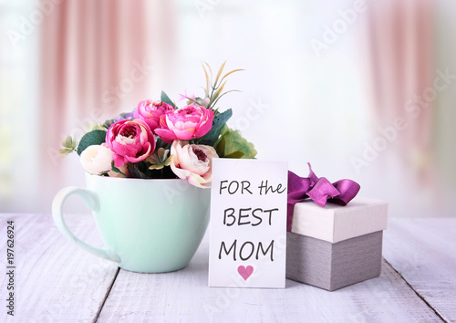Happy mothers day gifts with red white roses in a cup, and gift tag with text „For the best mom“, presented on wooden table with bright backround