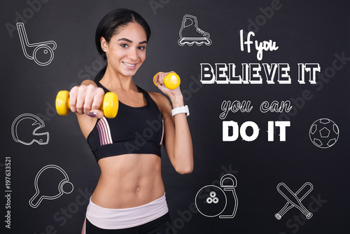 Strong person. Positive young active woman feeling strong and confident while being in a modern gym and doing exercises with yellow hand weights