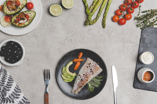 top view of fish fillet with baby carrots on plate and fresh healthy vegetables and fruits on grey
