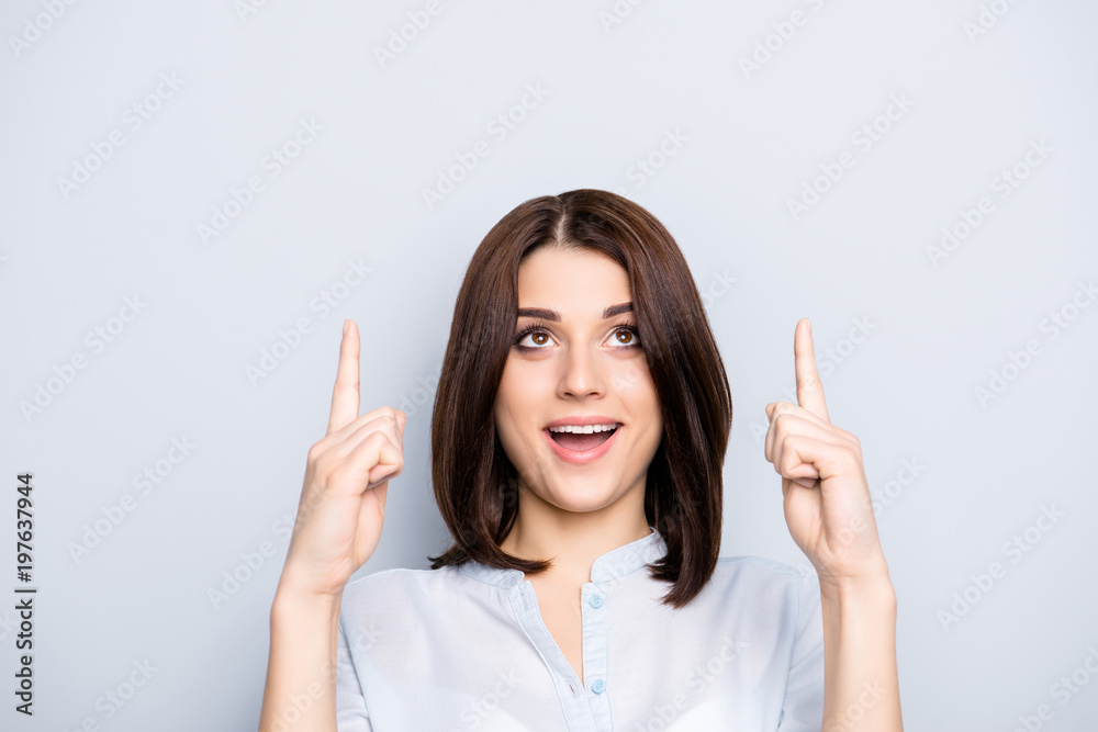 Close up portrait of charming, pretty, cheerful, stylish, trendy woman with modern hairstyle in shirt, pointing two forefingers up on empty place, looking at copy space, isolated on grey background Close up portrait of charming, pretty, cheerful, stylish, trendy woman with modern hairstyle in shirt, pointing two forefingers up on empty place, looking at copy space, isolated on grey background