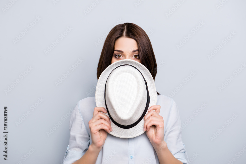 Oops, it is not me. Charming, pretty girl in shirt close her half face with hat, looking with mad eyes, hiding from someone, watching something, isolated on grey background