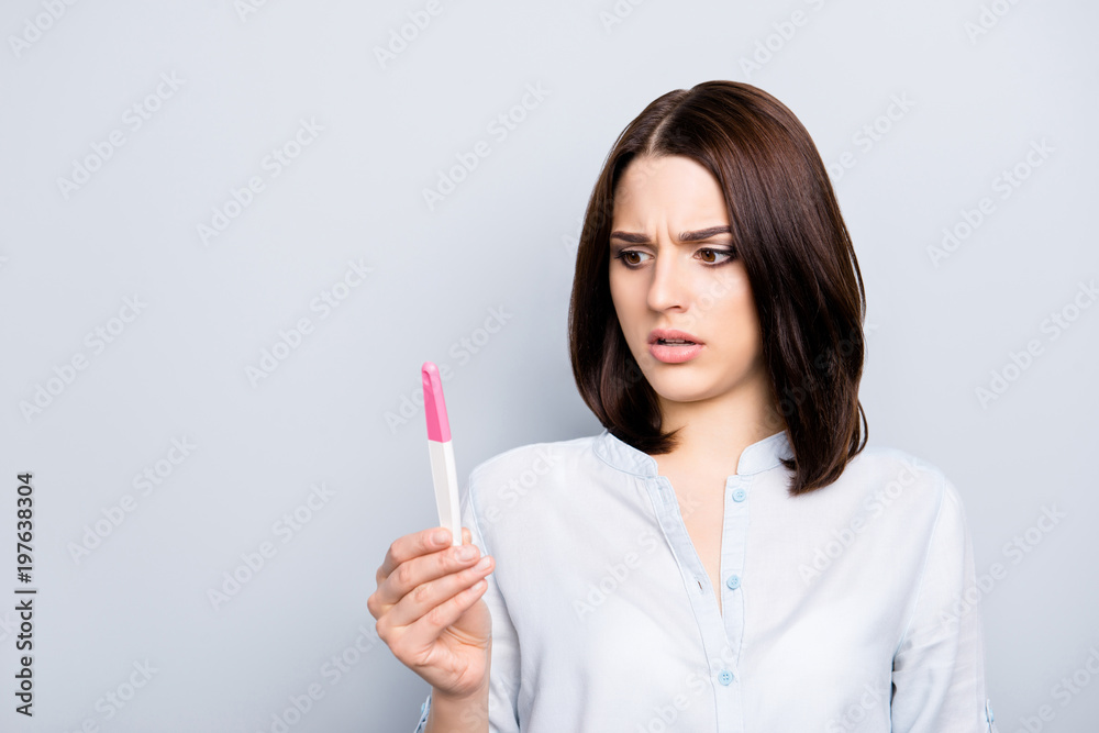 Unwished, unwanted, positive result fertility problem concept. Portrait of frustrated, upset, pretty, charming woman looking at pink and white tester in hand isolated on grey background Unwished, unwanted, positive result fertility problem concept. Portrait of frustrated, upset, pretty, charming woman looking at pink and white tester in hand isolated on grey background
