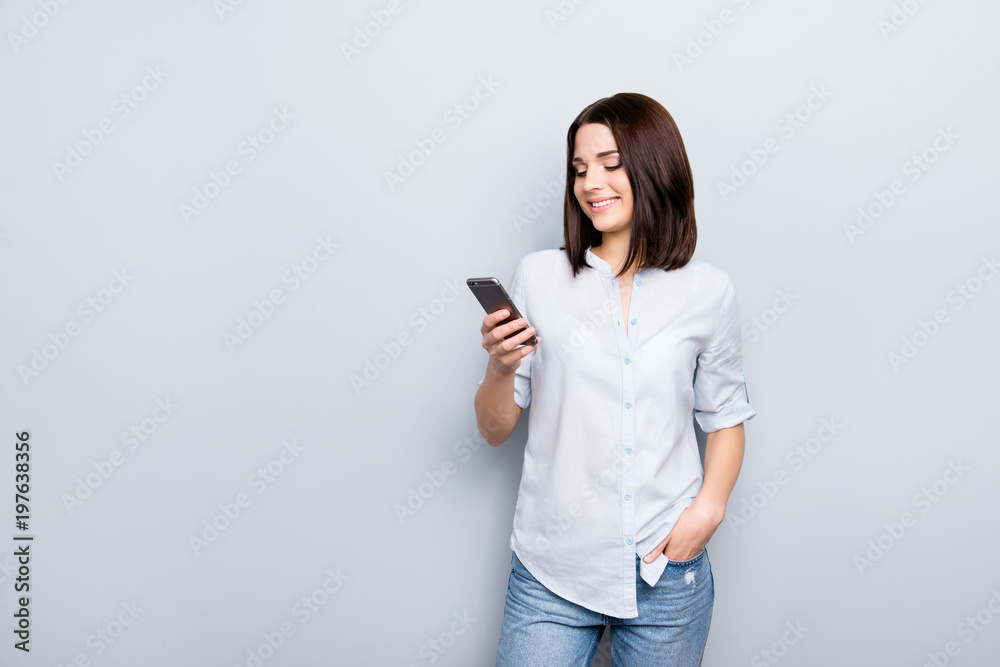 Portrait of pretty, charming, stylish, modern girl in shirt, denim outfit holding hand in pocket of jeans, having smart phone, checking email, searching, using 3G internet, standing on grey background Portrait of pretty, charming, stylish, modern girl in shirt, denim outfit holding hand in pocket of jeans, having smart phone, checking email, searching, using 3G internet, standing on grey background