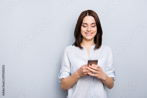 Portrait of pretty, charming, stylish, modern girl in denim outfit holding smart phone in hands, checking email, searching, using 5G internet, standing on grey background, chatting with lover, friends