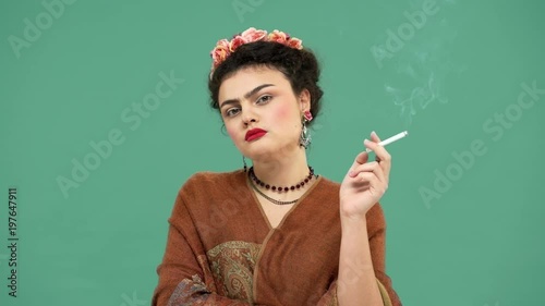 Portrait of elegant woman with thick eyebrows as Frida Kahlo with roses in hair looking seriously while smoking cigarette, isolated over green background. Stylization concept photo