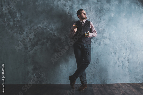 Man in vest standing, leaning against the wall, holding a glass. Full lenght portrait