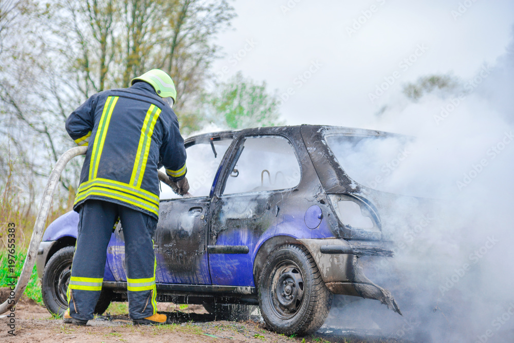 Obraz premium Fire fighter prepare to attack a propane fire. Burning and crashed car after explosion. Accident on street at countryside. No one was injured.