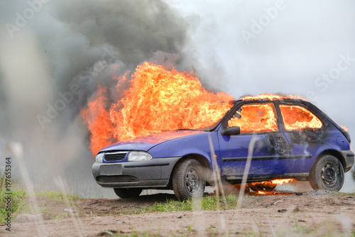 Burning and crashed car after explosion. Accident on street at countryside. Fire fighters prepare to attack a propane fire. No one is injured.