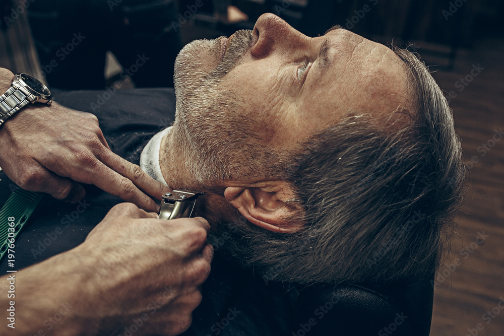 Naklejka premium Close-up side back view handsome senior bearded caucasian man getting beard grooming in modern barbershop.