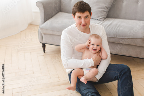 Adult man holding adorable laughing baby on hands sitting on floor near sofa smiling at camera