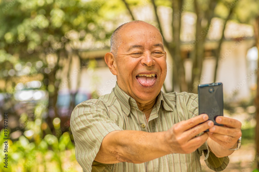 Obraz premium Outdoor view of old man sits on bench doing video cam sticking out tongue with cellphone in his hands, surrounding of nature. All problems left behind