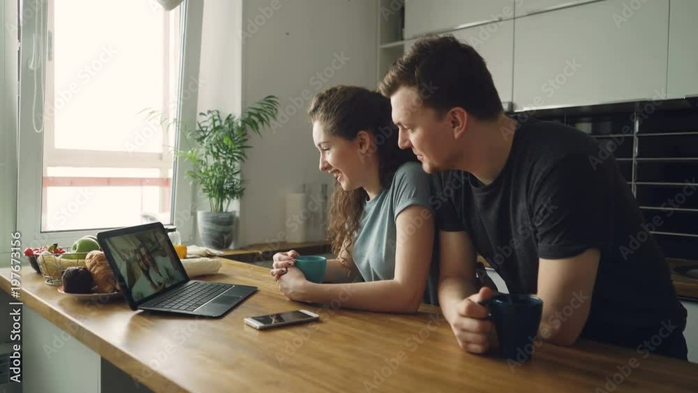 Young cheerful caucasian couple sitting at table in front of laptop skyping with their happy positive tanned friends from abroad, they are finishing conversation waving hands Young cheerful caucasian couple sitting at table in front of laptop skyping with their happy positive tanned friends from abroad, they are finishing conversation waving hands