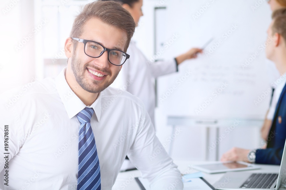 Business people at meeting in office. Focus at cheerful smiling bearded man wearing glasses. Conference, corporate training or brainstorming of people group. Success and negotiation concept Business people at meeting in office. Focus at cheerful smiling bearded man wearing glasses. Conference, corporate training or brainstorming of people group. Success and negotiation concept