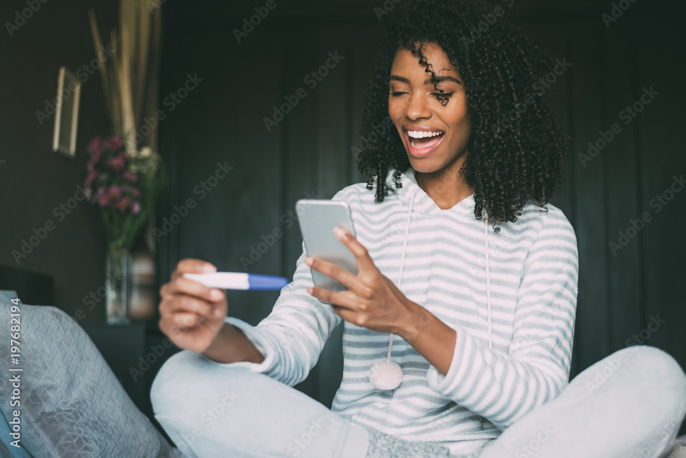 happy black woman with a pregnancy test and smartphone on bed happy black woman with a pregnancy test and smartphone on bed