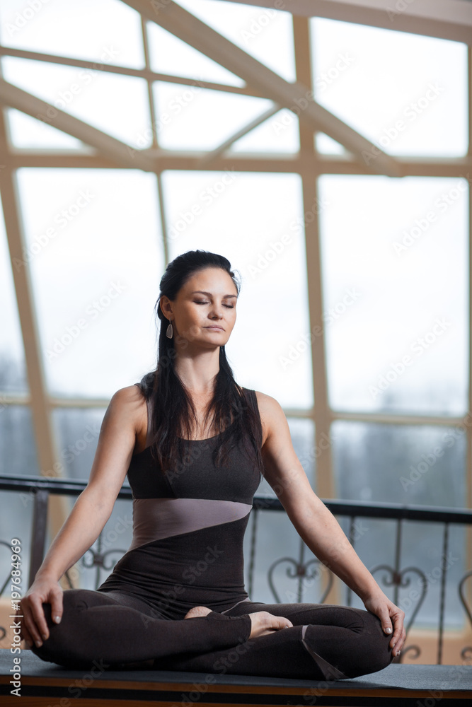 yogi woman with closed eyes in a dark jumpsuit practicing yoga meditating with serenity in front of the big window yogi woman with closed eyes in a dark jumpsuit practicing yoga meditating with serenity in front of the big window