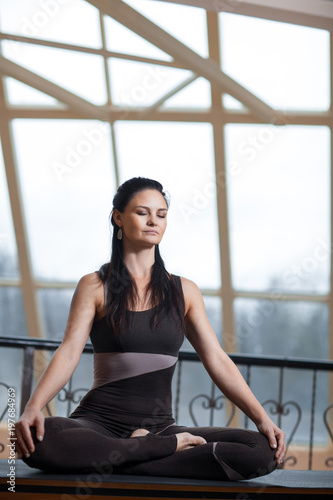 yogi woman with closed eyes in a dark jumpsuit practicing yoga meditating with serenity in front of the big window