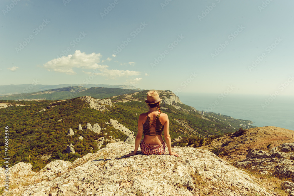 A beautiful young girl in a hat contemplates the beauty of mountains and watches nature. Far away in the background there is an azure sea, hills of mountains and greenery of dense forests.