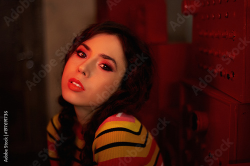 Studio fashion shooting. The girl in a bright outfit, top and pants flared in red and orange colors. Brunette with curly hair. Red light lamp in the twilight. The atmosphere of mystery