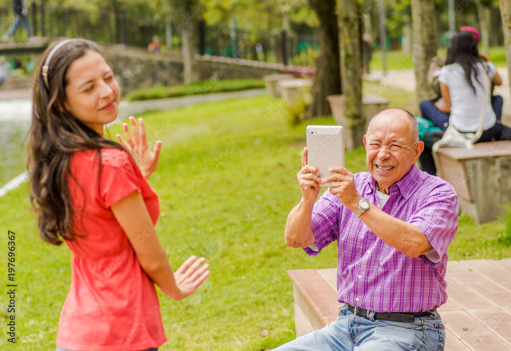 Fototapeta premium Outdoor view of father taking pictures with a tablet to his daughter while she is trying to avoid the pictures, in the park
