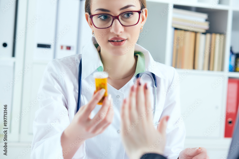 Young female medicine doctor in glasses sitting in front of working table holding jar of pills and writing prescription on special form. Young female medicine doctor in glasses sitting in front of working table holding jar of pills and writing prescription on special form.