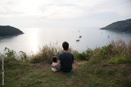 Daddy and his little daughter are relaxing. Baby girl with her father are sitting on a grass on the hill and looking to the sea during sunset.