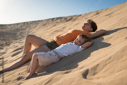 Mother and son relaxing on sand dune under blue sky