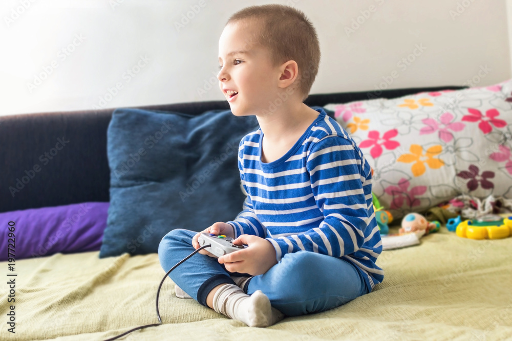 Cute boy playing video game console while sitting on sofa at home. Smiling and feeling happy