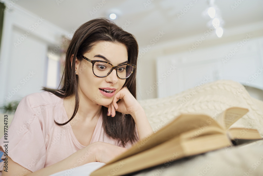 A girl in glasses lying on the sofa reading a book in the room. A girl in glasses lying on the sofa reading a book in the room.